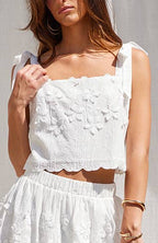 A woman models a white two-piece outfit consisting of a sleeveless crop top with floral embroidery and a matching flared skirt, paired with beige lace-up platform sandals, set against a white draped fabric backdrop.