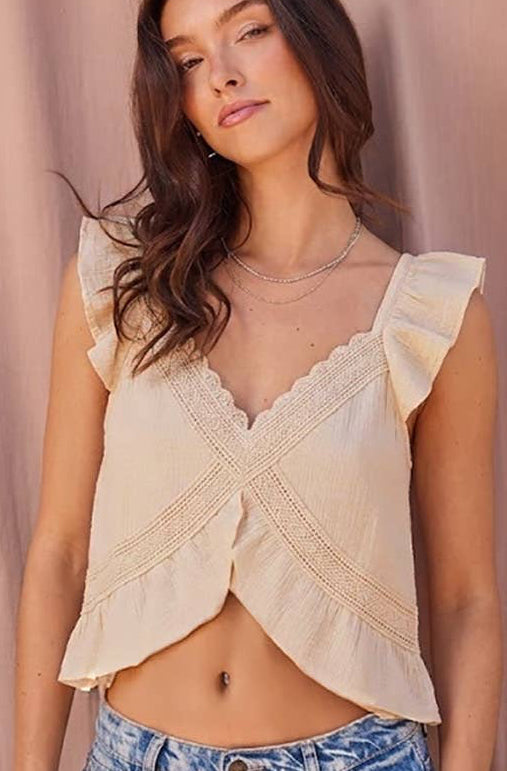 A woman wearing a cream ruffled crop top with lace trim and light wash blue jeans, standing against a draped dusty pink backdrop.