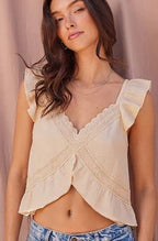 A woman wearing a cream ruffled crop top with lace trim and light wash blue jeans, standing against a draped dusty pink backdrop.