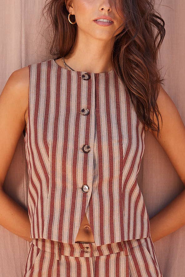 A woman wearing a sleeveless button-front top and matching striped pants, posed against a soft pink curtain backdrop.