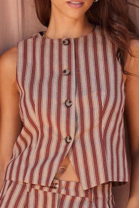 A woman modeling a sleeveless striped jumpsuit with a button-front bodice and wide-leg pants, posed against a draped dusty pink fabric backdrop.