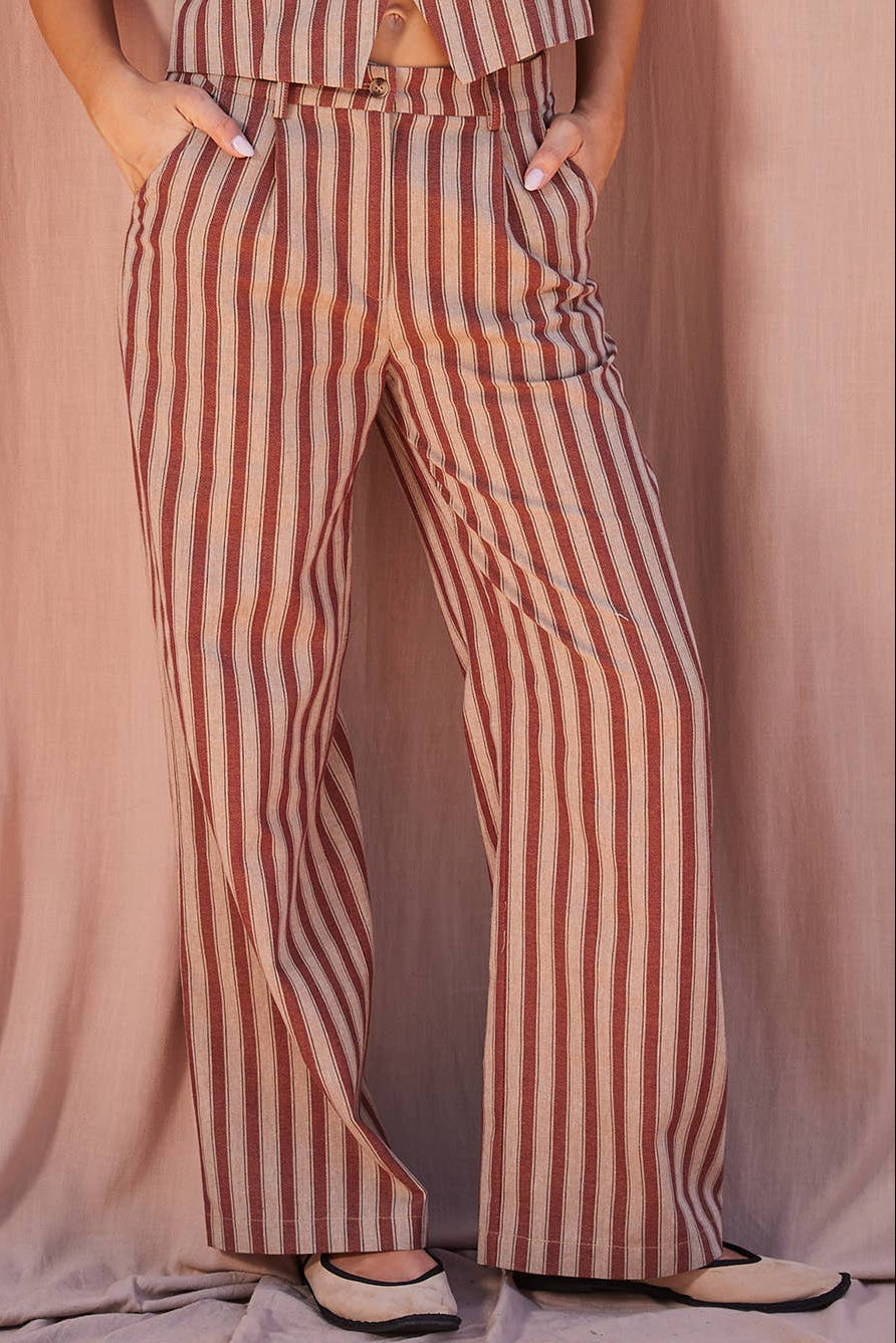 A person wearing high-waisted, wide-leg striped pants in vertical rust red and beige stripes, paired with a matching cropped top and beige flats, posed against a draped dusty pink fabric backdrop.
