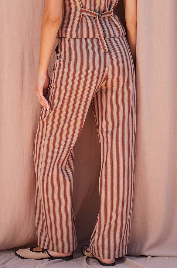 A woman stands with her back to the camera wearing a sleeveless wide-leg jumpsuit with vertical brown and beige stripes, set against a draped dusty pink fabric backdrop.