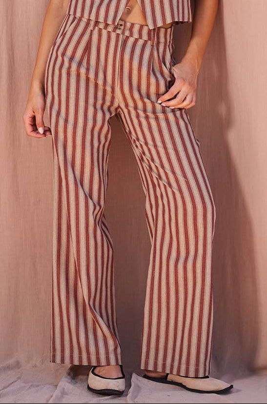 A woman models a sleeveless button-front top and matching wide-leg trousers in a vertical stripe pattern of rust red and beige, posed against a draped dusty pink fabric backdrop.