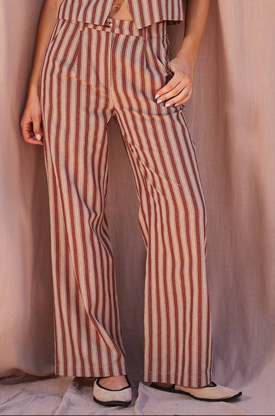A woman modeling a sleeveless striped jumpsuit with a button-front bodice and wide-leg pants, posed against a draped dusty pink fabric backdrop.