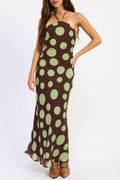 A woman modeling a brown halter-neck maxi dress with a large light green polka dot pattern, paired with brown leather boots, against a plain white studio background.