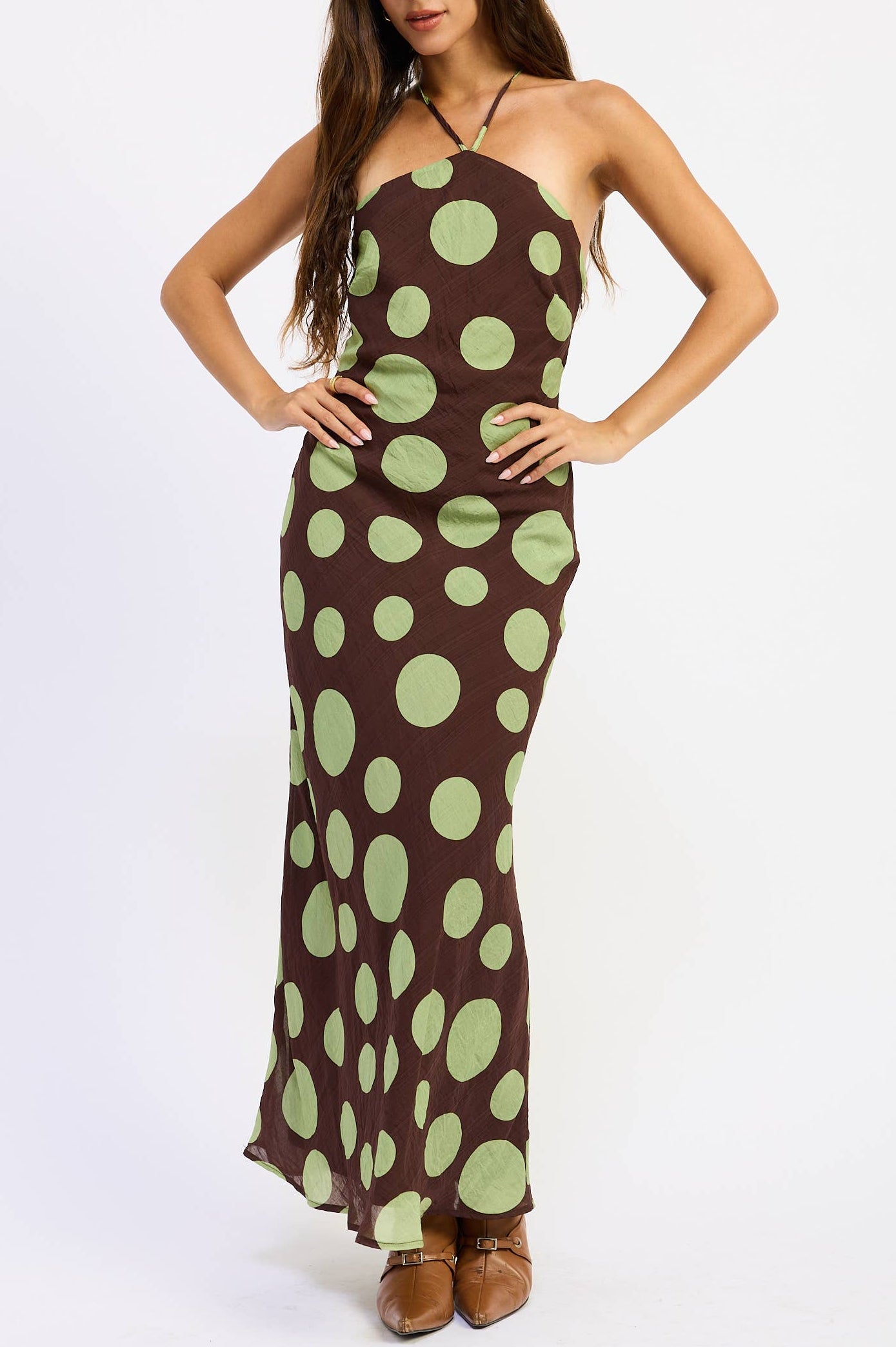 A woman modeling a brown halter-neck maxi dress with a large light green polka dot pattern, paired with brown shoes, against a plain white studio background.