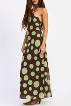 A woman modeling a one-shoulder maxi dress with a brown base and large light green polka dots, paired with brown heeled shoes, against a plain white studio background.