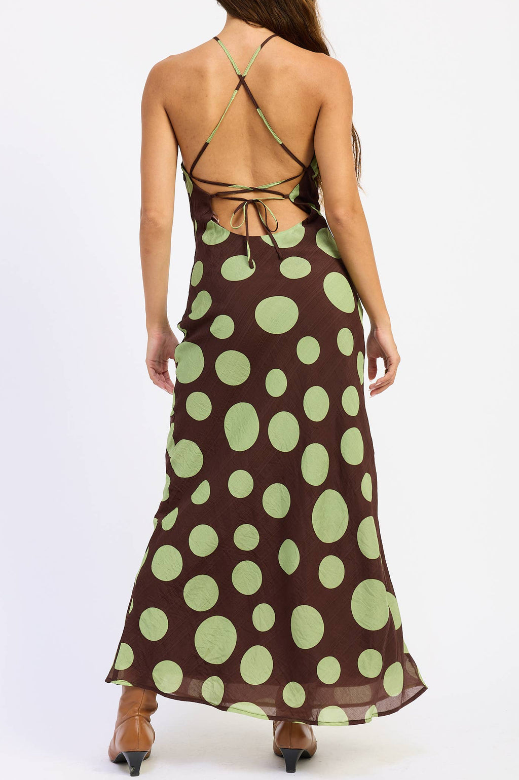 A woman viewed from the back wearing a brown halter maxi dress with large light green polka dots and an open back, standing against a plain white studio background.