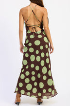A woman viewed from the back wearing a brown halter maxi dress with large light green polka dots and an open back, standing against a plain white studio background.