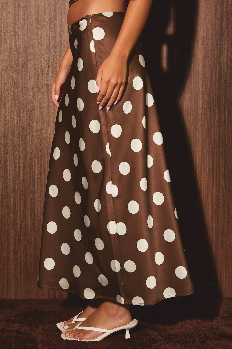A model wearing a brown satin maxi skirt with large white polka dots, paired with a textured crop top and white heeled sandals, standing against a dark wood-paneled wall.