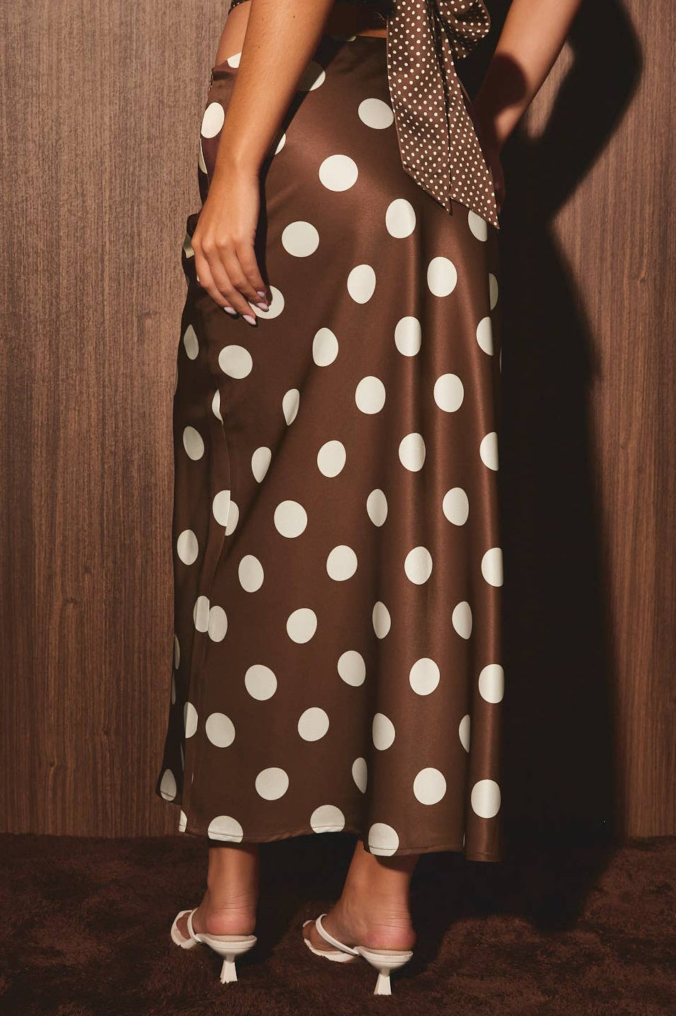 A woman wearing a brown satin maxi skirt with large white polka dots, paired with a matching polka-dot tie belt and white kitten-heel sandals, standing against a dark wood-paneled wall.