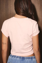 A woman viewed from behind wearing a light pink short-sleeve crop top and blue denim jeans, standing against a dark wood-paneled background.