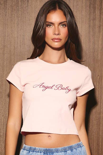 A woman stands against a brown wood-paneled wall wearing a light pink cropped t-shirt with 'Angel Baby' printed in dark script, a light blue denim mini skirt, and white strappy heels.