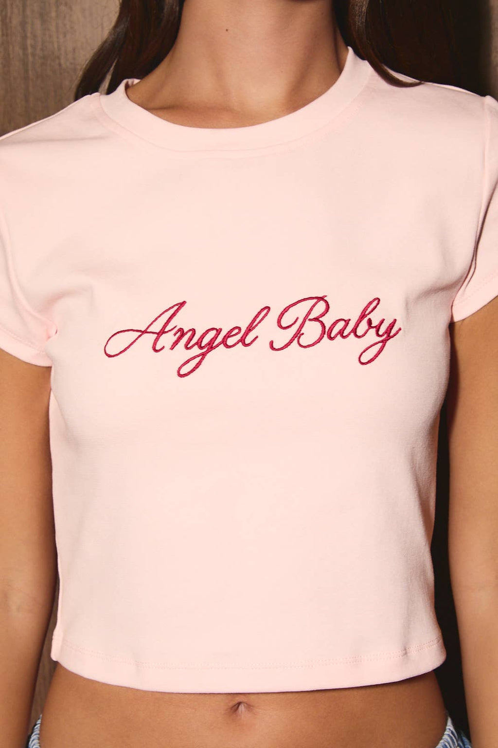A close-up of a model wearing a light pink short-sleeve crop top with 'Angel Baby' written across the chest in red cursive script.