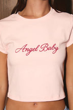 A close-up of a model wearing a light pink short-sleeve crop top with 'Angel Baby' written across the chest in red cursive script.