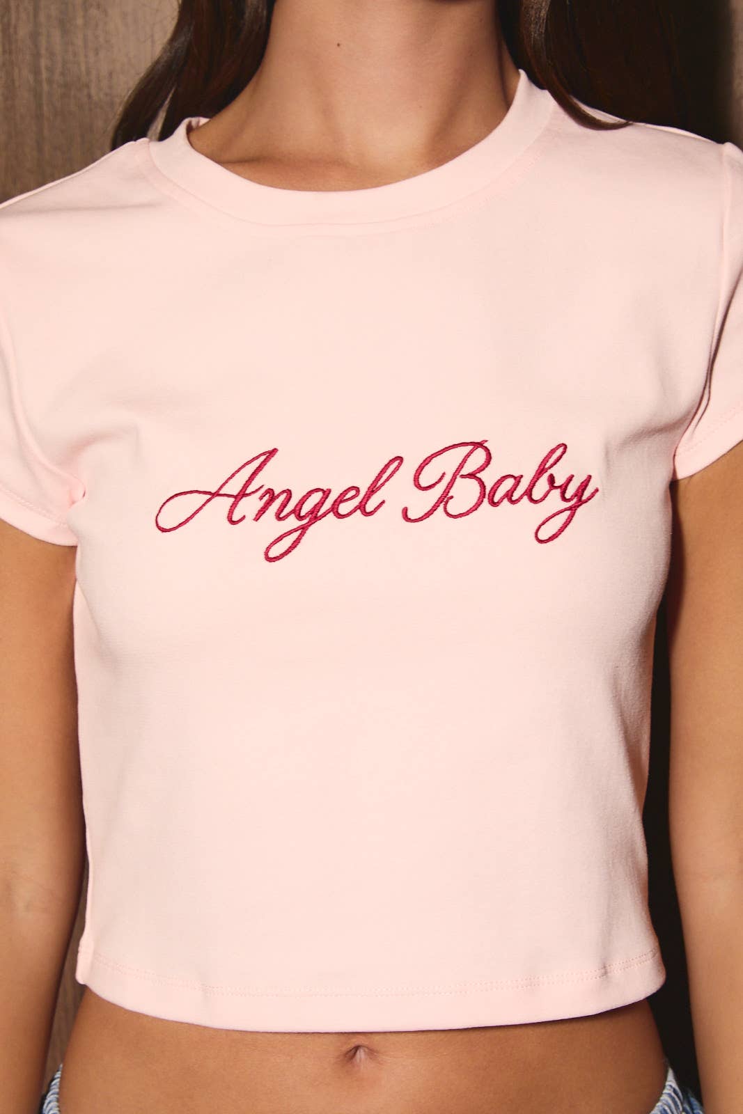 A close-up of a model wearing a light pink short-sleeve crop top with 'Angel Baby' written across the chest in red cursive script.