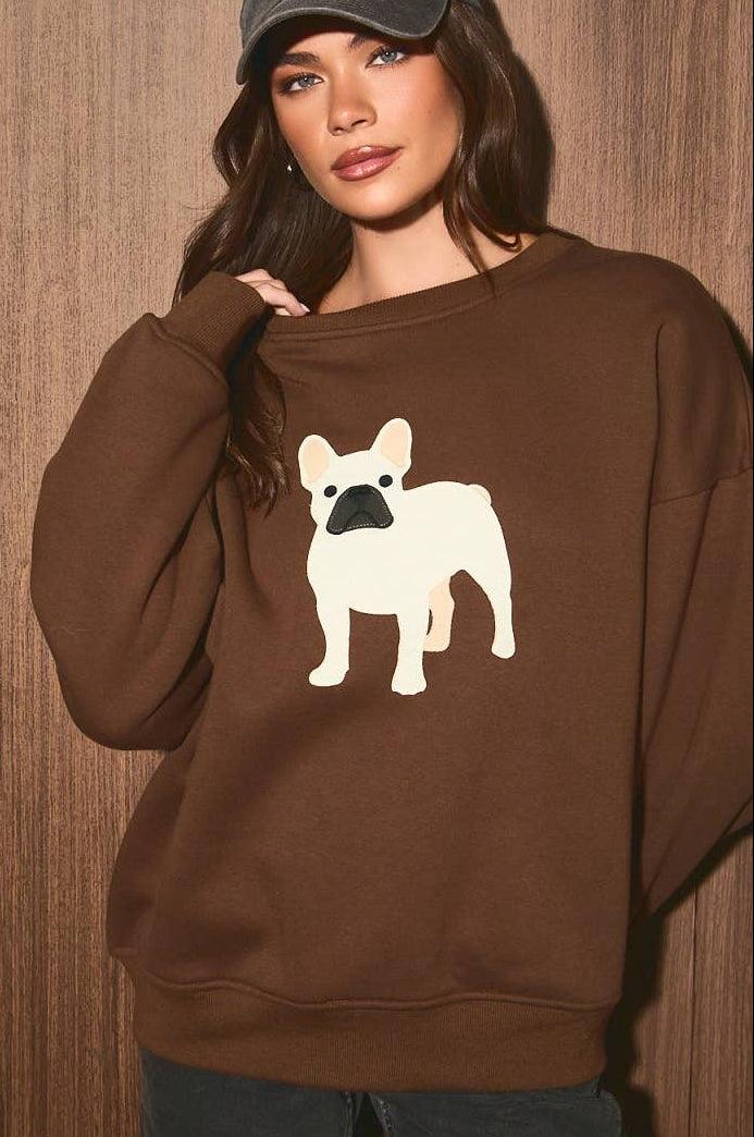 A woman wearing a brown sweatshirt with a white French Bulldog graphic on the front, paired with a dark gray baseball cap and dark jeans, standing against a brown wood-paneled background.