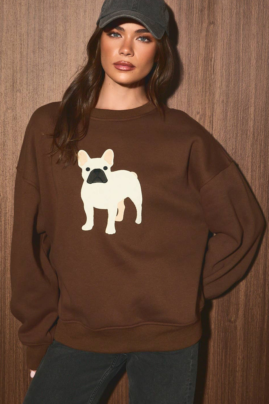 A woman wearing a brown crewneck sweatshirt with a white French Bulldog graphic on the front, paired with a dark gray baseball cap and dark jeans, standing against a brown wood-paneled background.