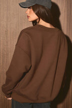 A woman wearing an oversized brown crewneck sweatshirt, a dark gray baseball cap, and dark blue jeans, standing against a textured brown wooden wall.