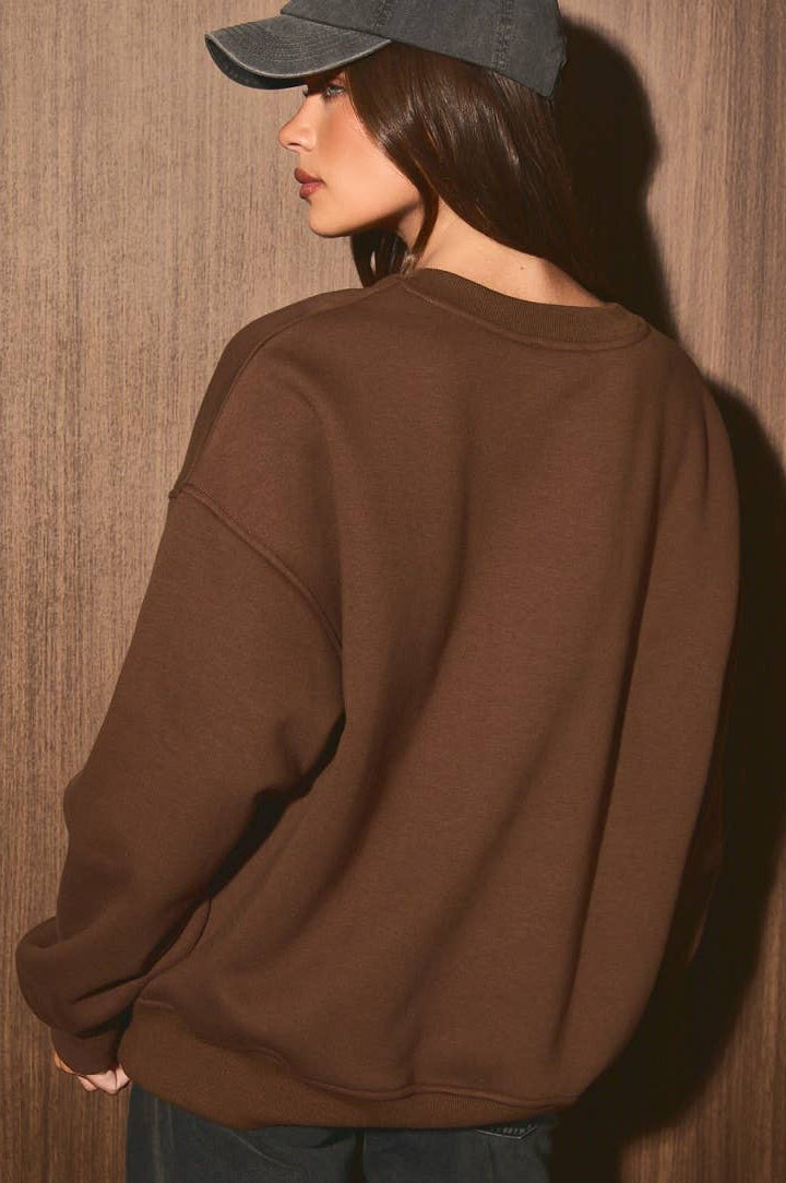 A woman wearing an oversized brown crewneck sweatshirt, a dark gray baseball cap, and dark blue jeans, standing against a textured brown wooden wall.