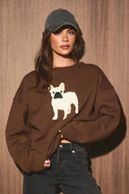 A woman stands against a brown wood-paneled wall wearing a brown sweatshirt with a white French bulldog graphic, dark wide-leg jeans, tan slip-on shoes, and a dark gray baseball cap.