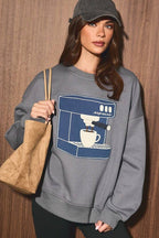 A woman wearing a gray crewneck sweatshirt with a blue and white coffee machine graphic, black bike shorts, a dark gray baseball cap, and carrying a tan suede tote bag, standing against a brown wood-paneled wall.