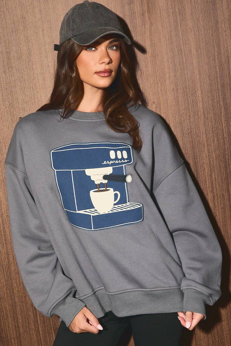 A woman wearing a gray crewneck sweatshirt with a blue coffee machine and cup graphic on the front, paired with black bike shorts and a dark gray cap, standing against a brown wood-paneled wall.