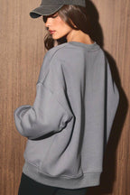 A woman wearing an oversized light gray sweatshirt, black bike shorts, and a dark gray baseball cap, standing against a brown wood-paneled wall.