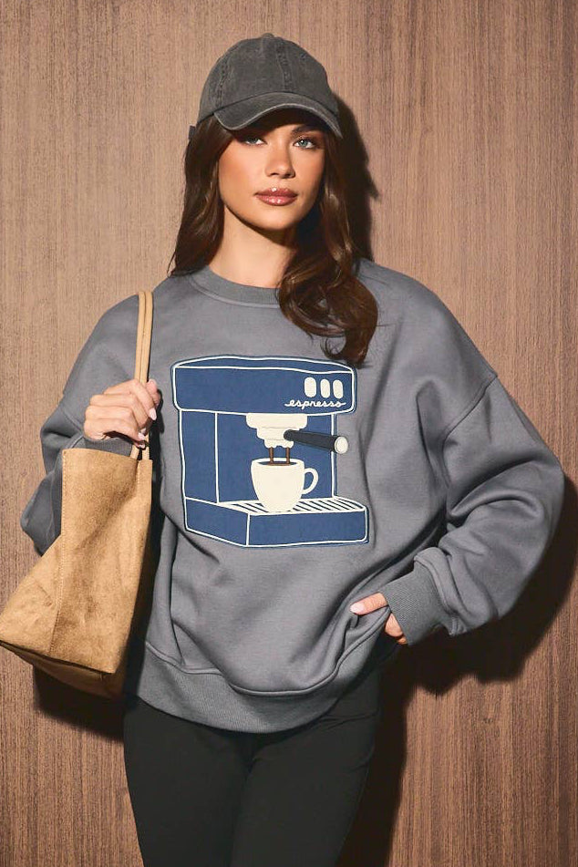 A woman stands against a wood-paneled wall wearing a gray sweatshirt with a large blue and white coffee machine graphic on the front, black capri leggings, black shoes, and carrying a tan shoulder bag.