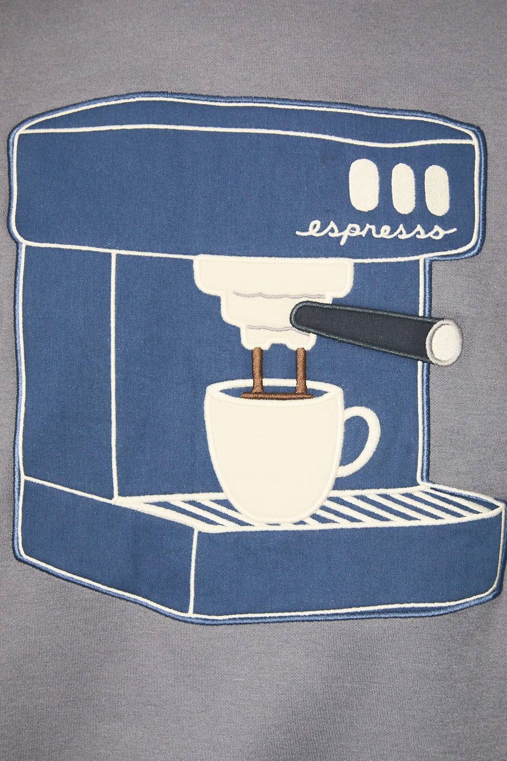 A close-up of a gray fabric garment featuring a large blue and white embroidered patch depicting an espresso machine with a cup and the word 'espresss'.