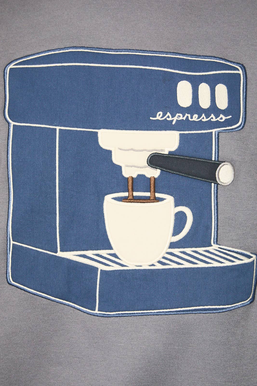 A close-up of a gray fabric garment featuring a large blue and white embroidered patch depicting an espresso machine with a cup and the word 'espresss'.