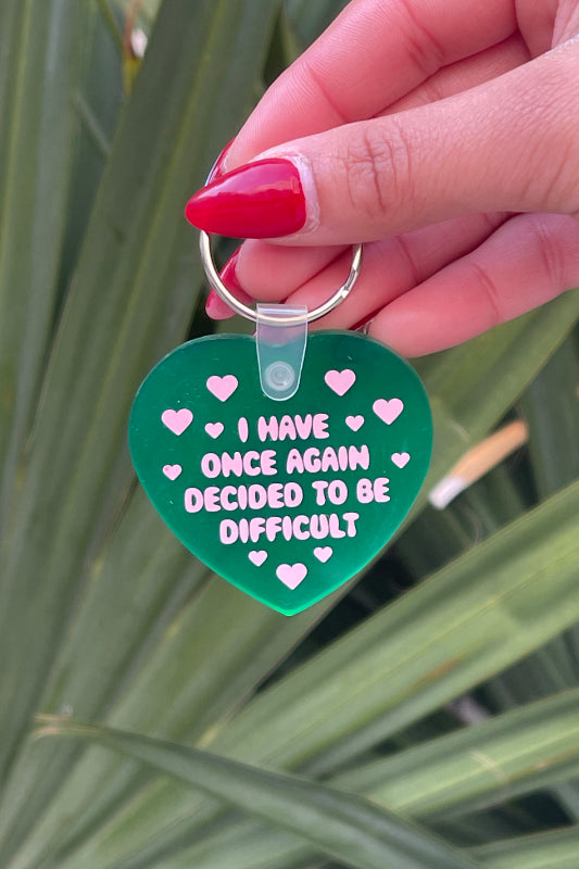 Green heart-shaped keychain with text held by a hand against a leafy background