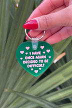 Green heart-shaped keychain with text held by a hand against a leafy background