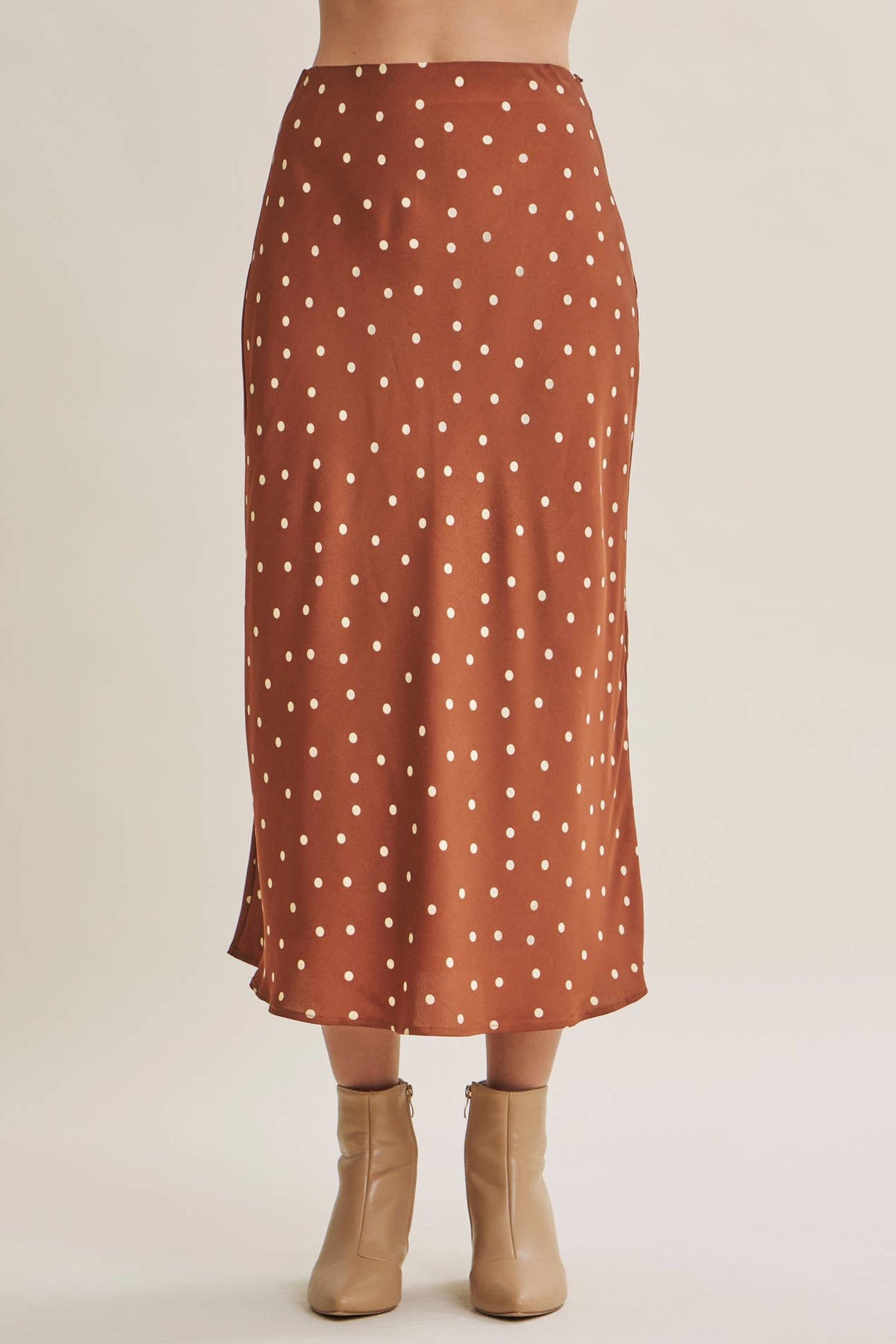 A rust-orange midi skirt with a white polka-dot pattern, shown on a model from the waist down wearing beige ankle boots against a light beige studio background.