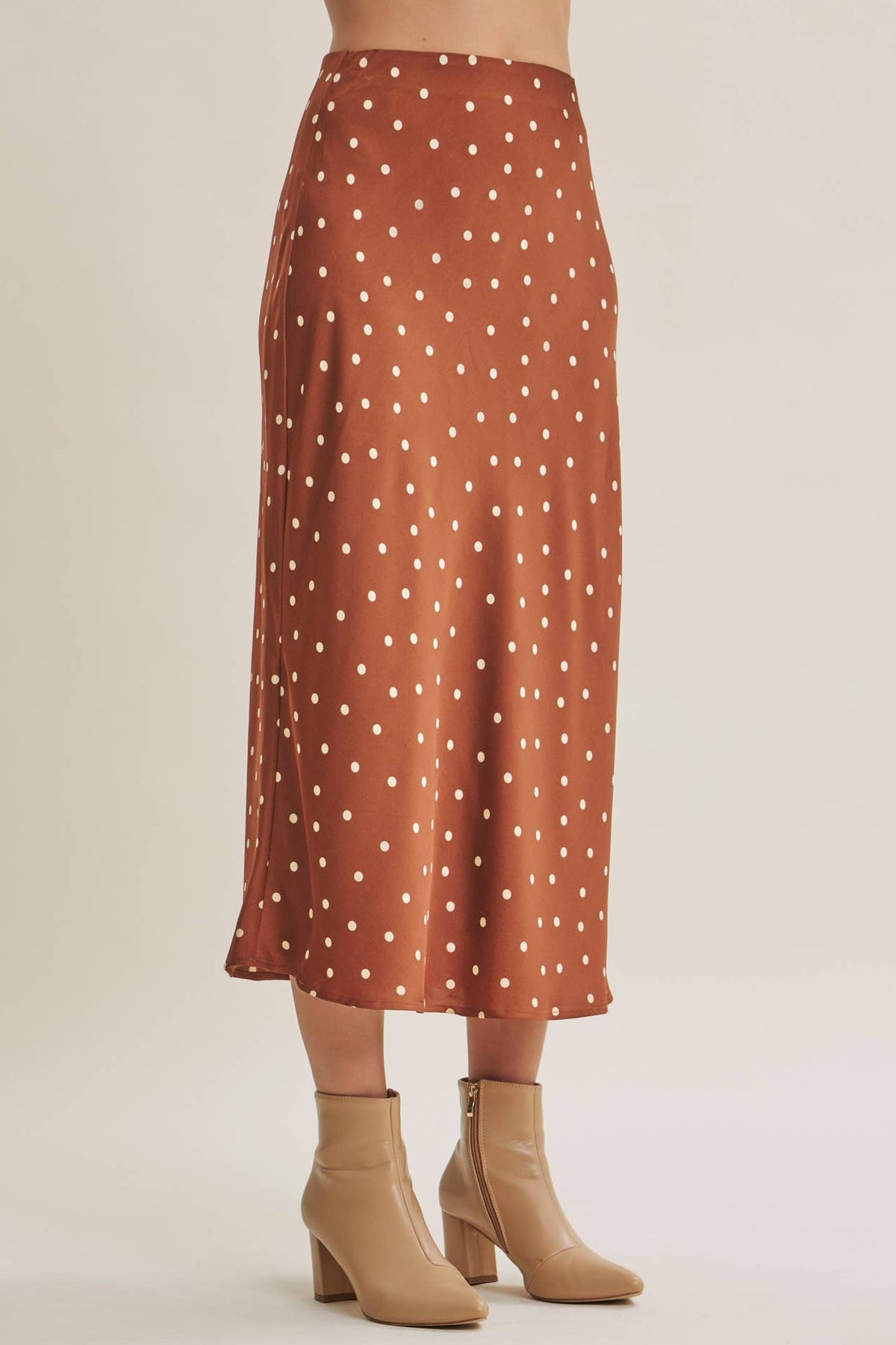A model is shown from the waist down wearing a rust-orange satin midi skirt with white polka dots, paired with beige ankle boots against a light beige studio background.
