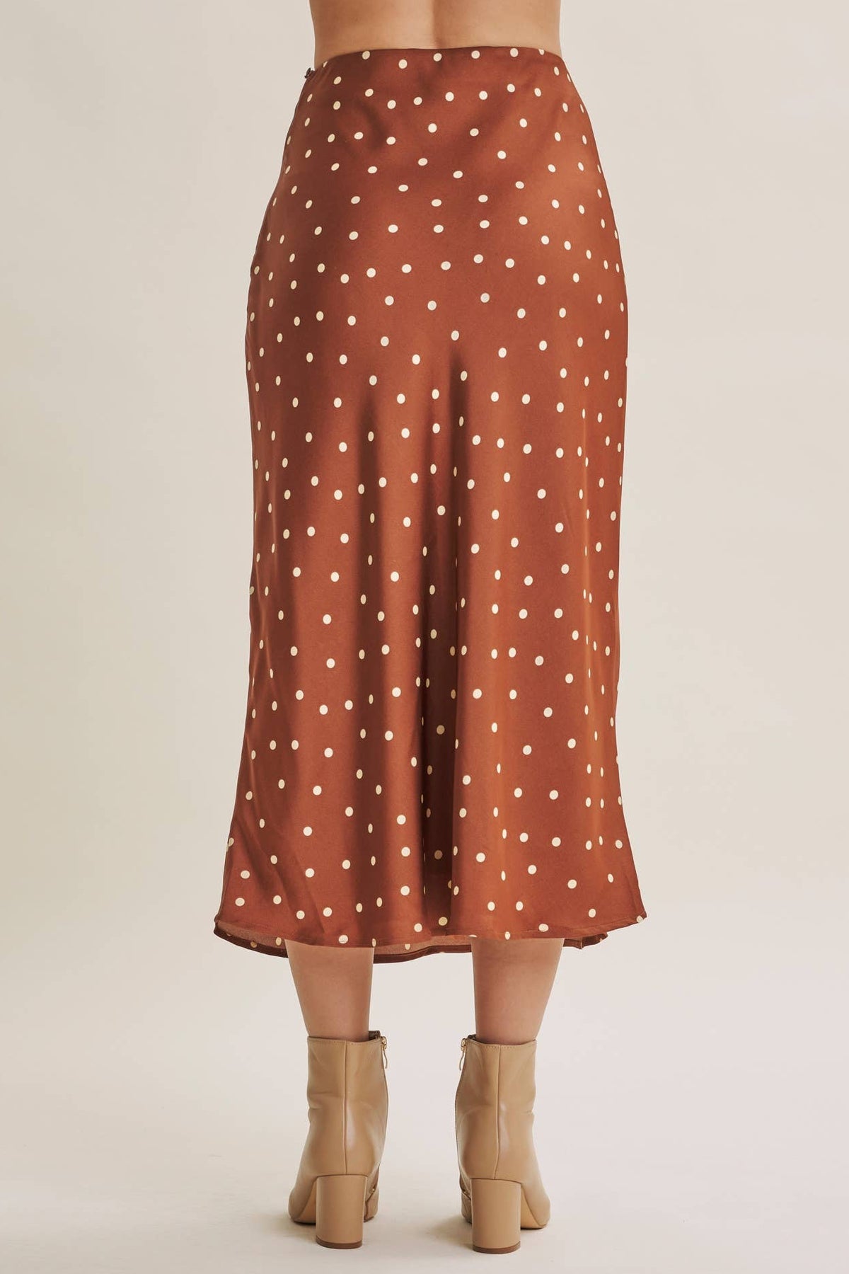 A model from the back wearing a rust-brown midi skirt with white polka dots and beige ankle boots, set against a plain beige studio background.