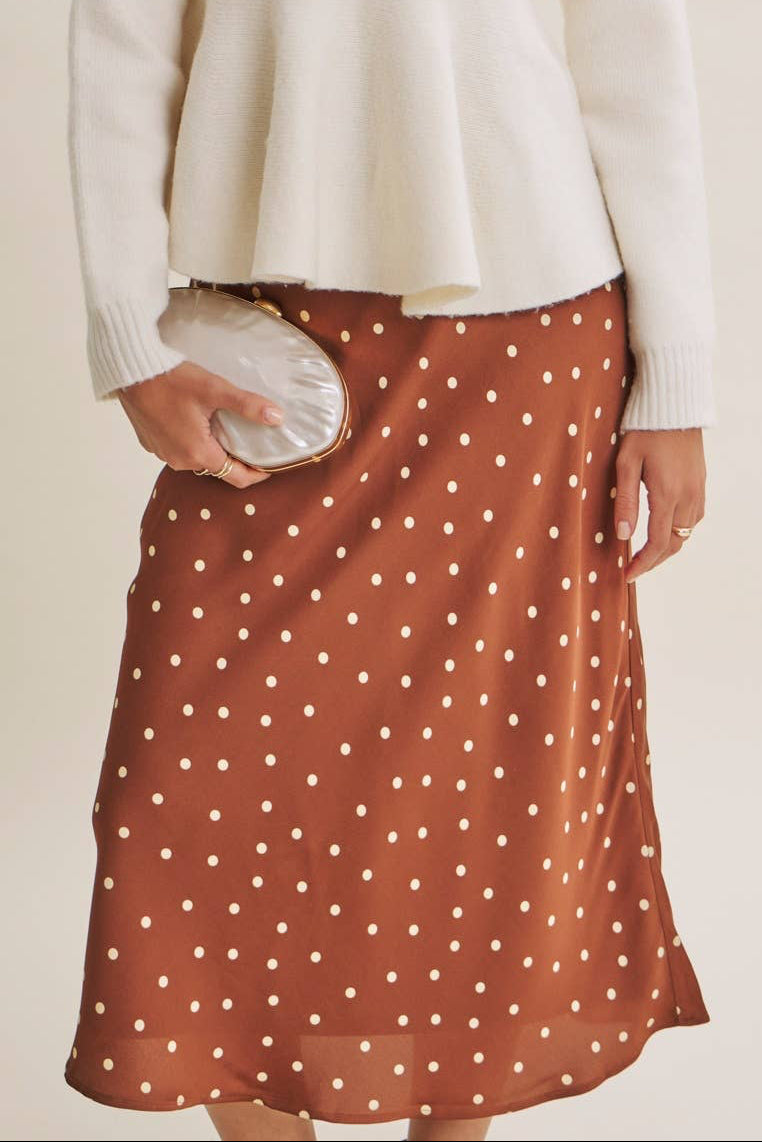 A woman modeling a cream long-sleeve knit top and a rust-orange midi skirt with white polka dots, holding a small metallic clutch against a plain beige studio background.