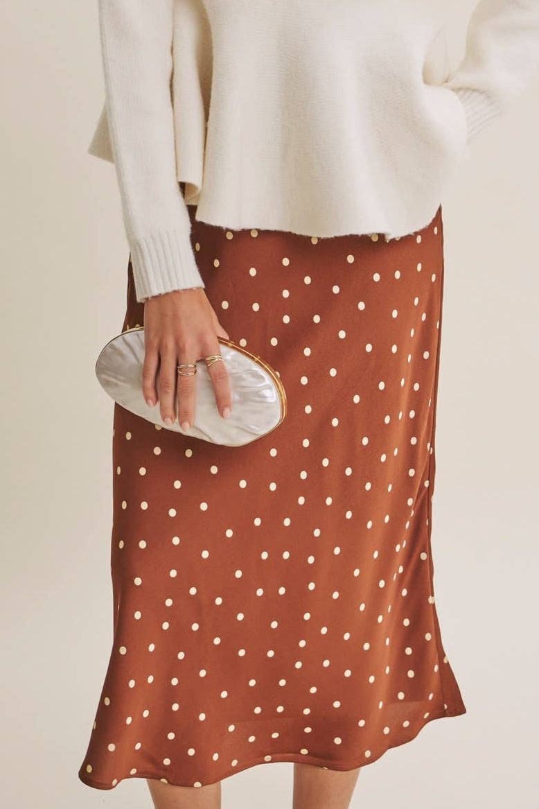A woman modeling a cream knit sweater and a rust-brown midi skirt with white polka dots, holding a white clutch against a plain beige studio background.