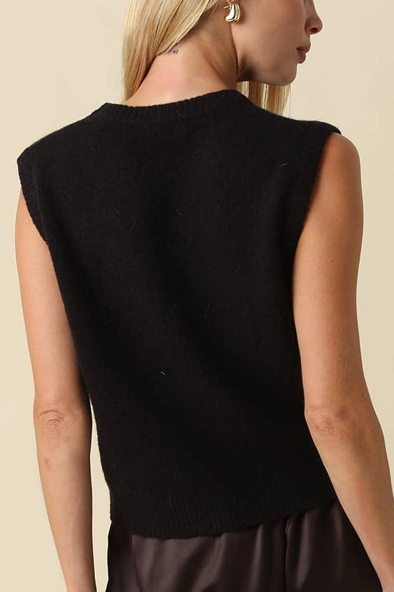 A woman shown from the back wearing a sleeveless black knit top and dark brown pants against a plain beige studio background.