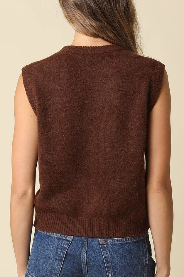 A woman viewed from behind wearing a sleeveless brown knit sweater vest and blue denim shorts against a plain beige studio background.