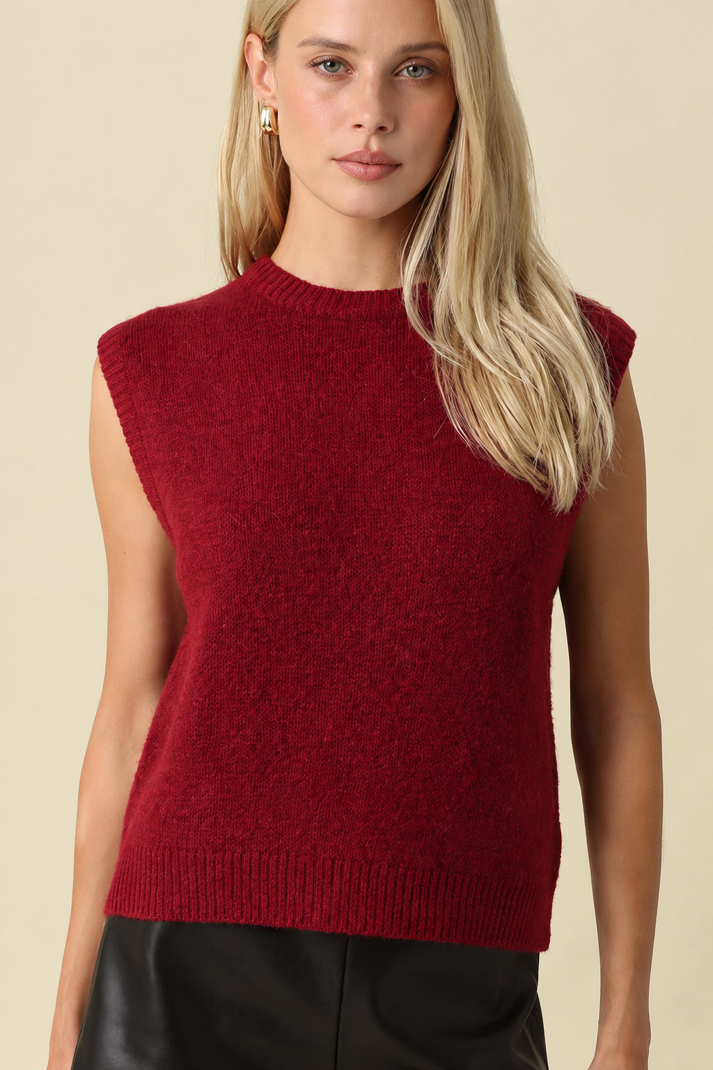 A blonde woman wearing a sleeveless, crewneck knit sweater vest in deep burgundy red, paired with black faux-leather shorts, against a solid beige background.