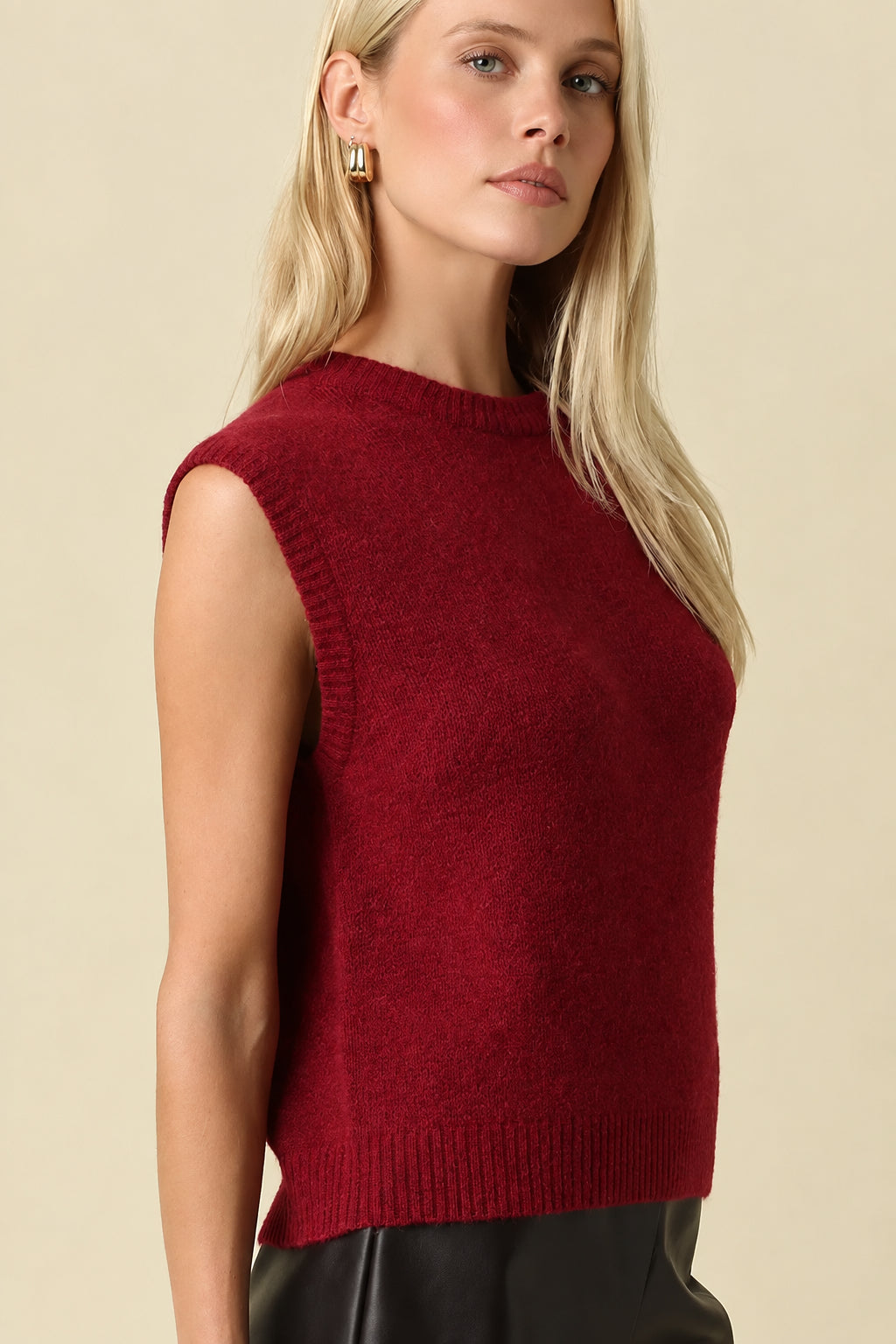 A blonde woman wearing a sleeveless, crew-neck knit sweater in deep burgundy red, paired with black faux-leather shorts, standing against a solid beige background.