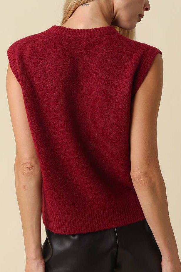 A woman shown from the back wearing a sleeveless, crew-neck knit sweater in deep burgundy, paired with black leather shorts, against a solid beige background.