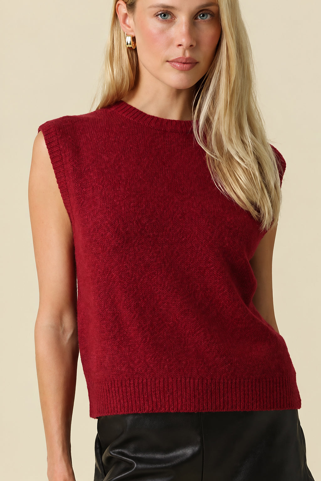 A blonde model wearing a sleeveless textured burgundy knit top, black shorts, and black cowboy boots, standing against a solid beige studio background.
