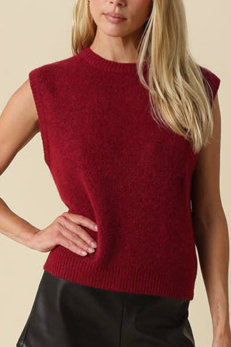 A blonde model wearing a sleeveless textured burgundy knit top, black faux-leather shorts, and black high-heeled boots, standing against a solid beige studio background.