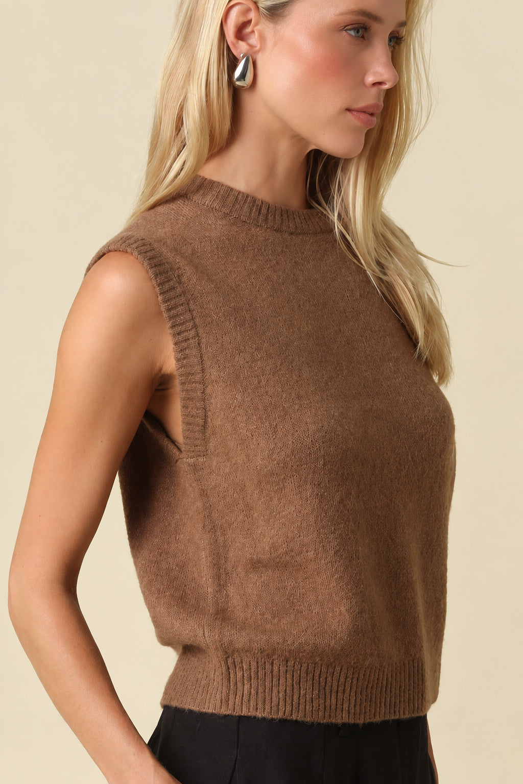 A blonde woman wearing a sleeveless brown knit sweater vest and black trousers, photographed against a solid beige studio background.
