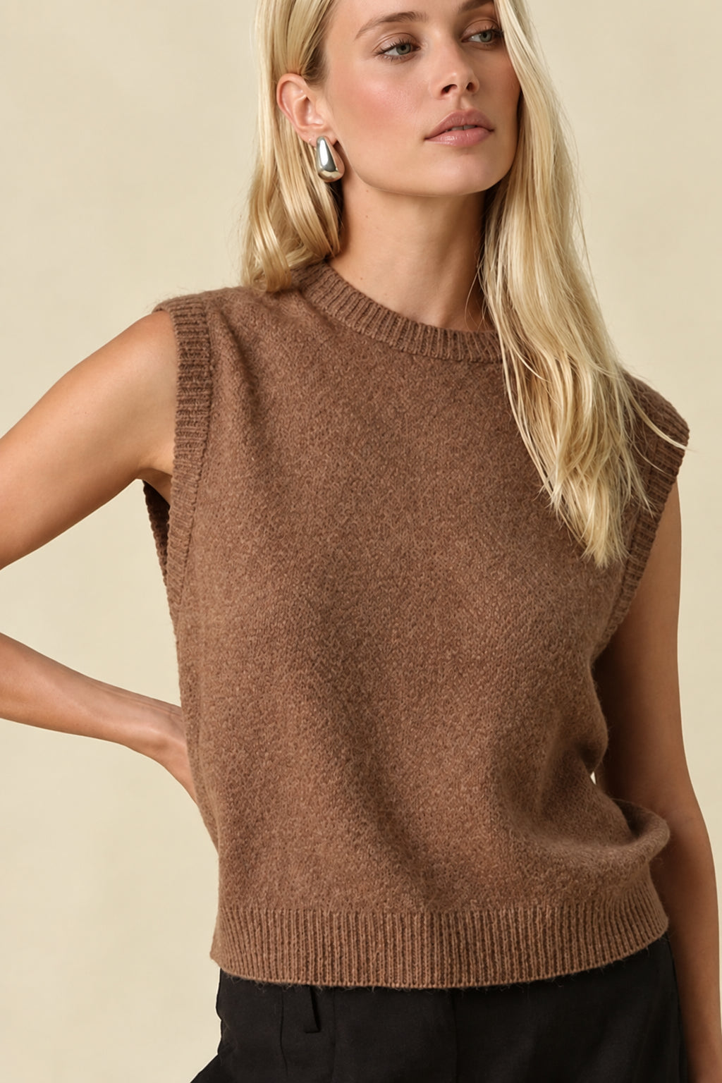 A woman modeling a sleeveless brown knit sweater vest paired with black shorts and black Mary Jane flats, standing against a solid beige studio background.