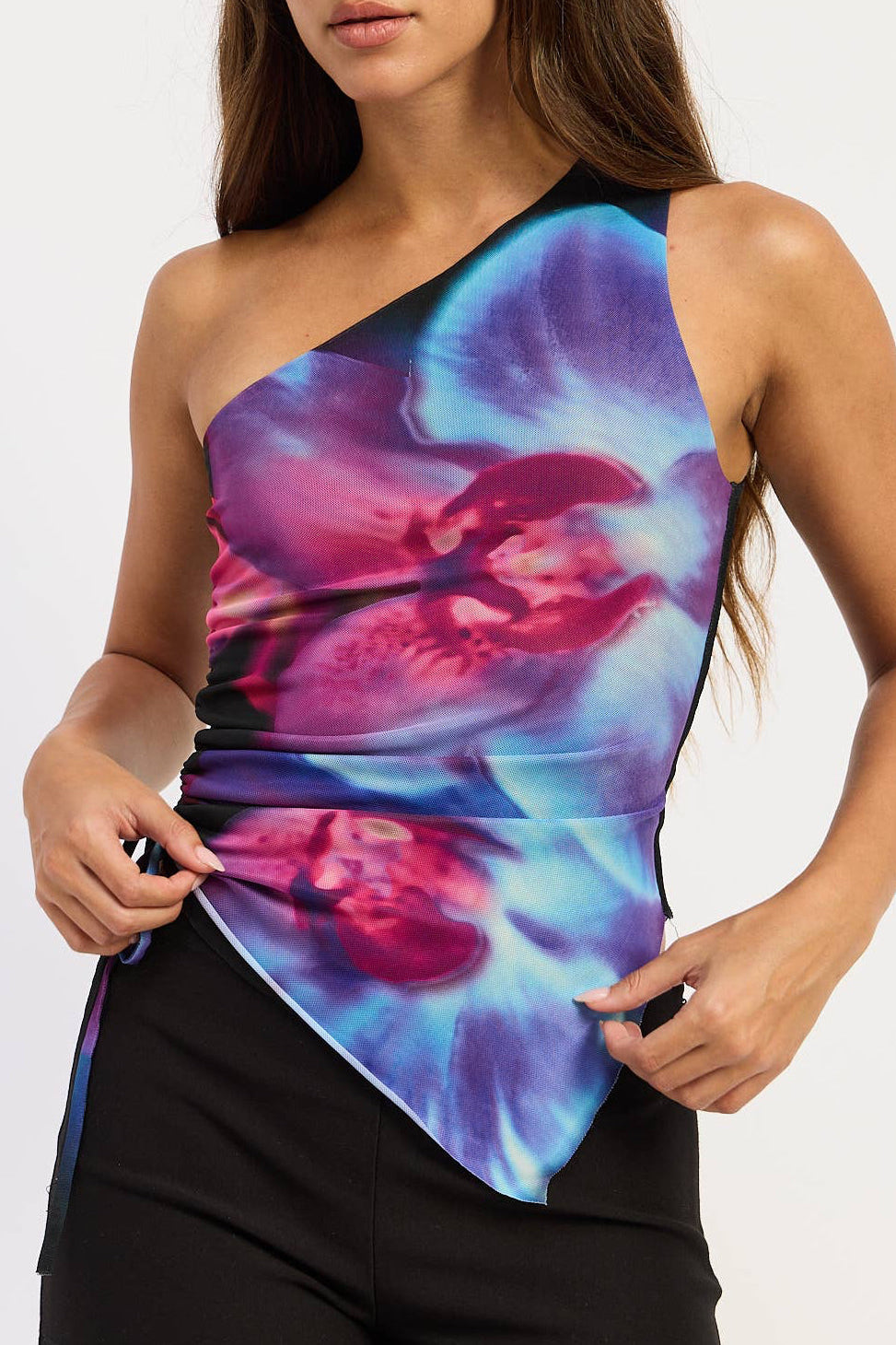 A woman modeling a one-shoulder athletic top with a vibrant blue, purple, and pink abstract print, paired with black athletic shorts, against a plain white background.