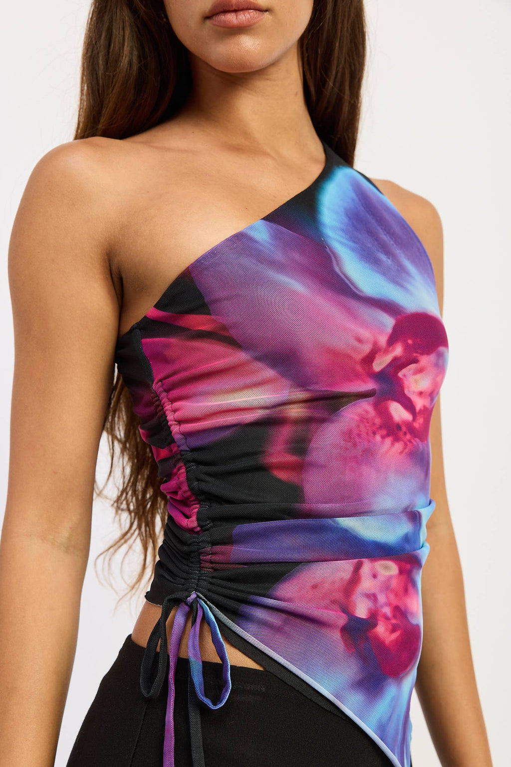 A close-up of a model wearing a one-shoulder ruched top with a vibrant purple, pink, and blue abstract floral print over a black mesh base.